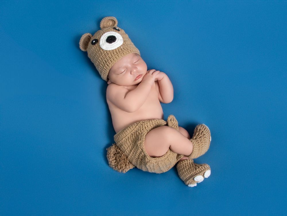 Sleeping newborn baby wearing a knitted teddy bear hat and outfit on a blue background.