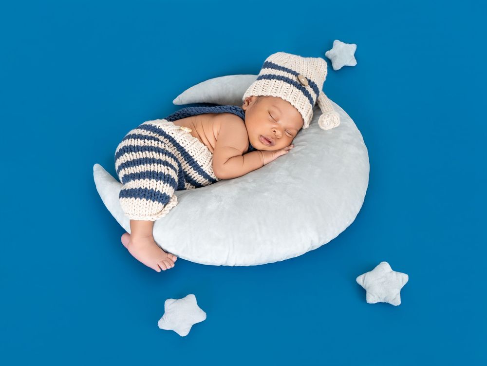 Newborn baby sleeping peacefully on a white crescent moon pillow, wearing striped knit pants and a star-tipped hat.