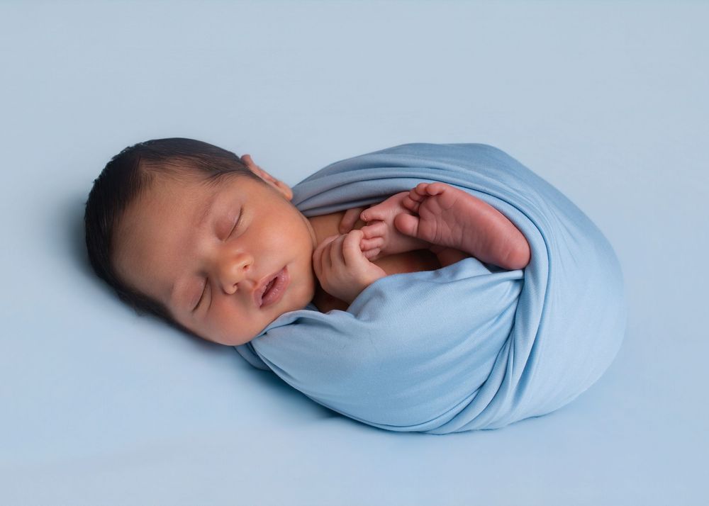 Sleeping newborn baby swaddled in a soft blue wrap, posed on a light blue background in a professional photo.