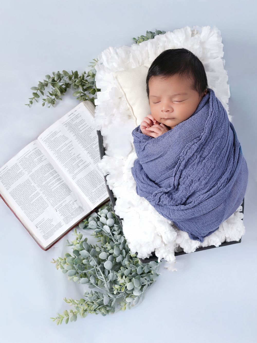 Sleeping newborn swaddled in blue wrap beside an open Bible and eucalyptus greenery on white florals.