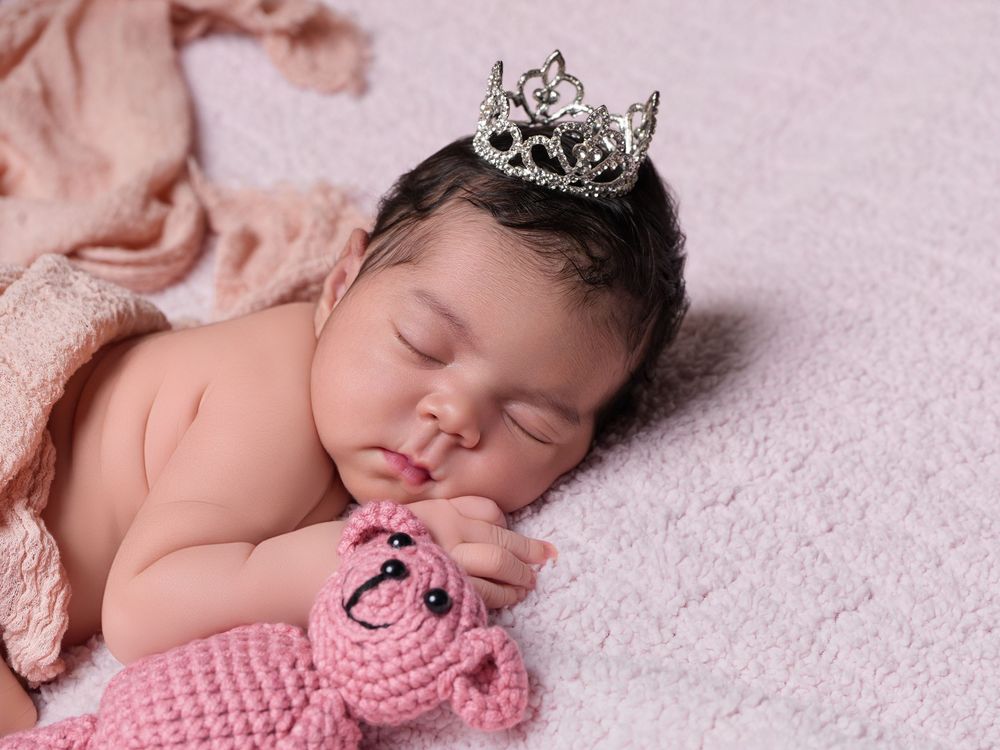 Sleeping newborn baby wearing a silver tiara crown, holding a pink crocheted teddy bear on a soft white blanket.