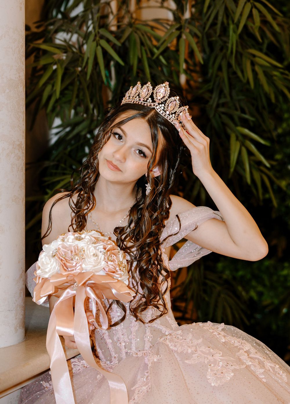 Young Latina girl in pink quinceañera gown holding bouquet and adjusting jeweled crown with curly hair.
