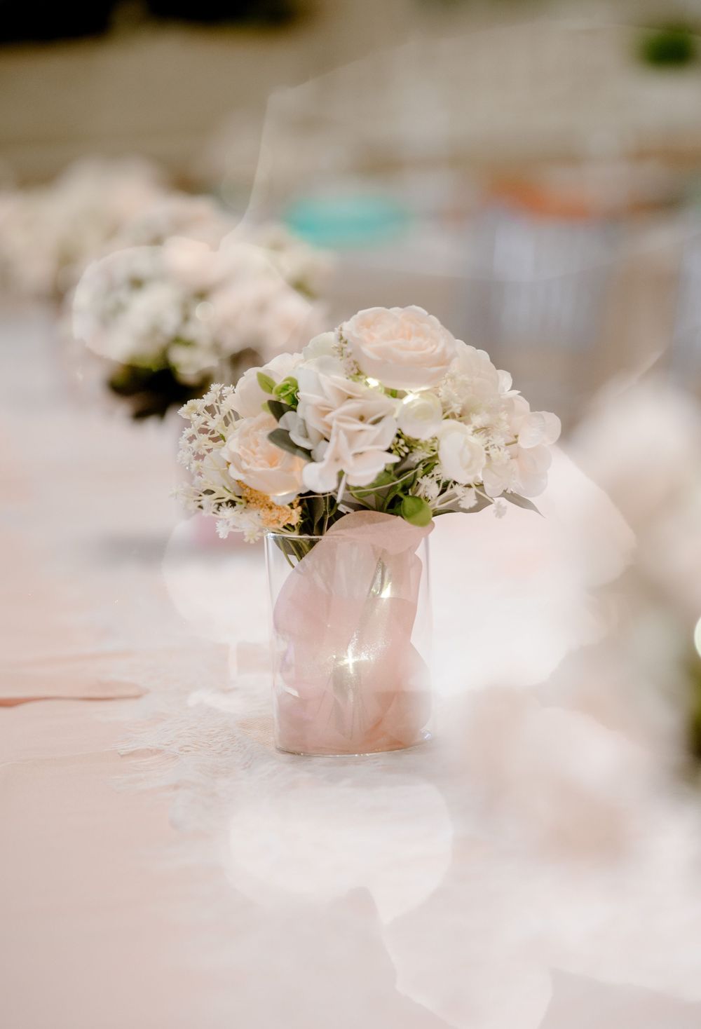 Elegant blush pink floral centerpiece with white roses in twisted vase on white tablecloth.