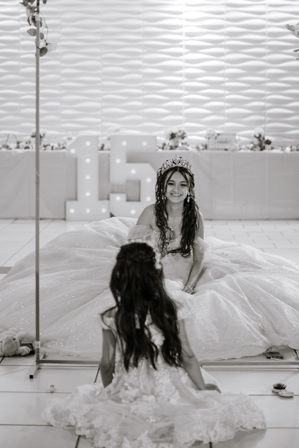 Black and white photo of a smiling quinceañera in a tiara and ball gown sitting on a dance floor.