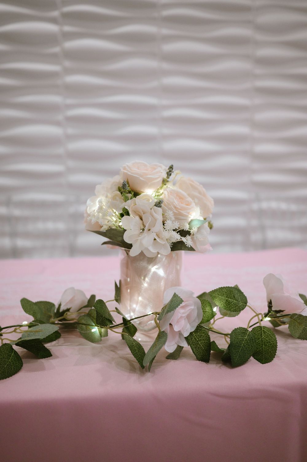 Pink roses and white flowers in a glowing mason jar vase with eucalyptus garland on a pink tablecloth.