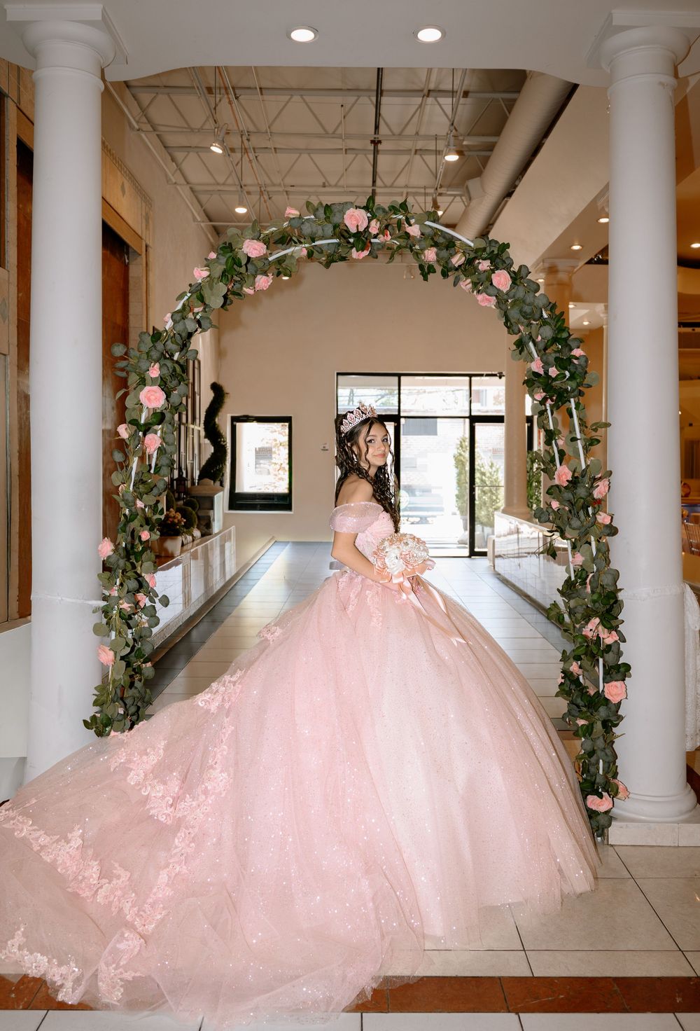 Young woman in pink quinceañera ballgown poses under floral arch with roses and greenery in elegant venue.