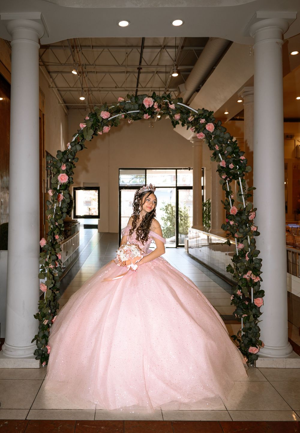 Young woman in pink ball gown quinceañera dress poses under floral arch with roses at elegant venue.