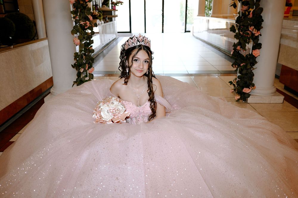Young girl in pink sparkly quinceañera ball gown with tiara holding bouquet in elegant floral hallway.