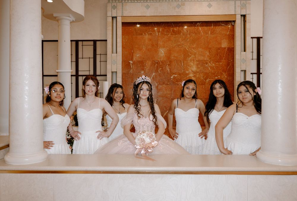 Seven young women in white gowns pose together, with the quinceañera wearing a tiara and pink bouquet at center.