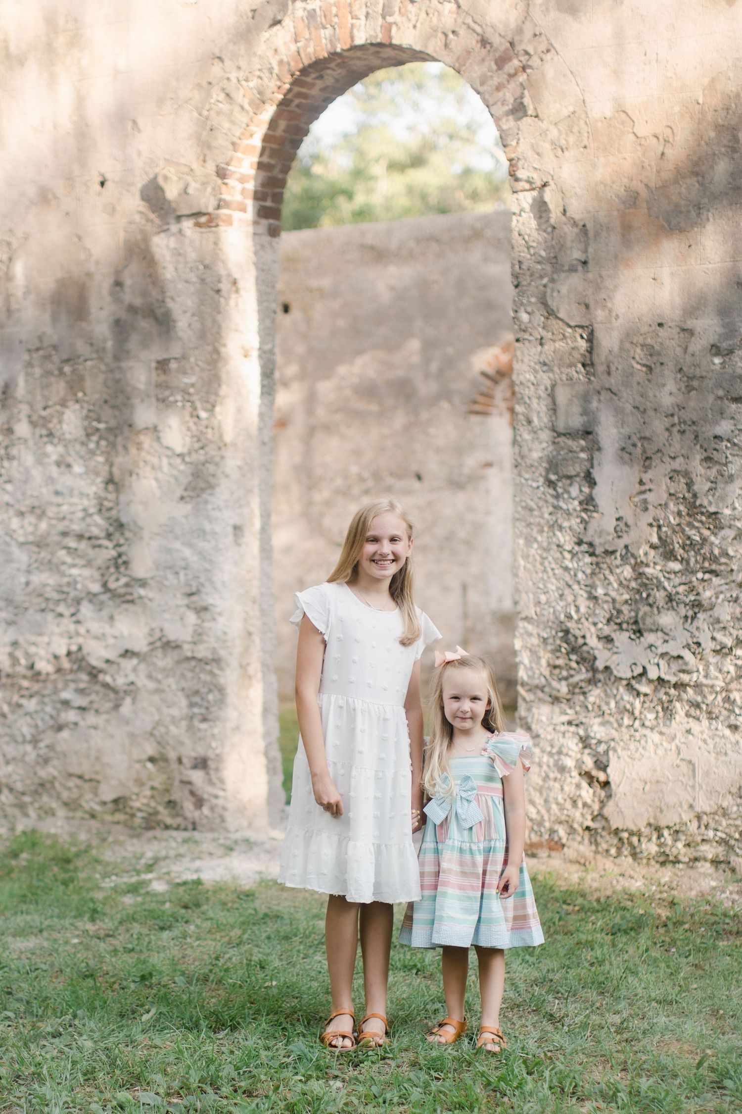 Two young girls in St. Helena, SC dressed in dresses smile before historic stone ruins with an arched doorway on a sunny day.