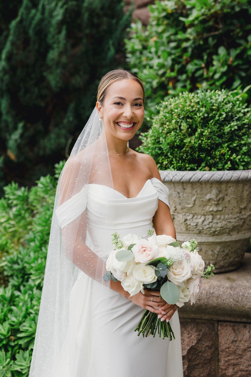Smiling bride in white off-shoulder gown and veil holds bouquet of blush roses outdoors near stone planter. Ocean Cliff Newport RI