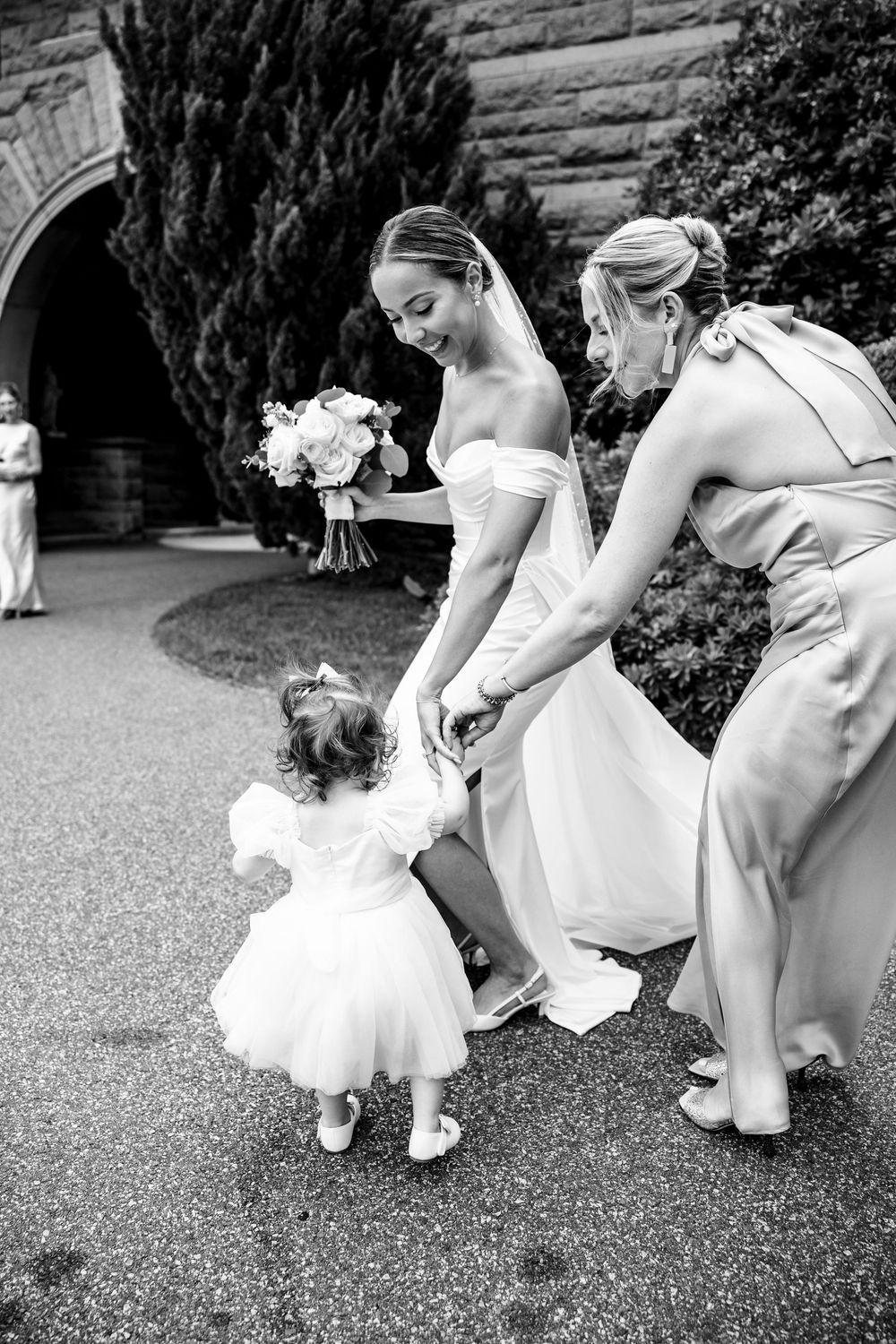 Black and white photo of a bride and bridesmaid bending down to greet a flower girl outside . OceanCliff Newport RI Wedding