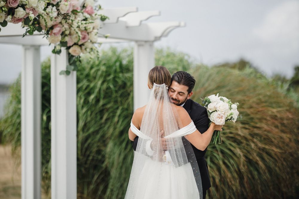 A bride and groom share a romantic embrace under a white floral pergola at an outdoor wedding ceremony.