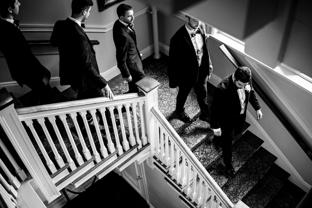 Groomsmen in tuxedos descend an elegant staircase in a black and white wedding photo. Ocean Cliff Newport RI