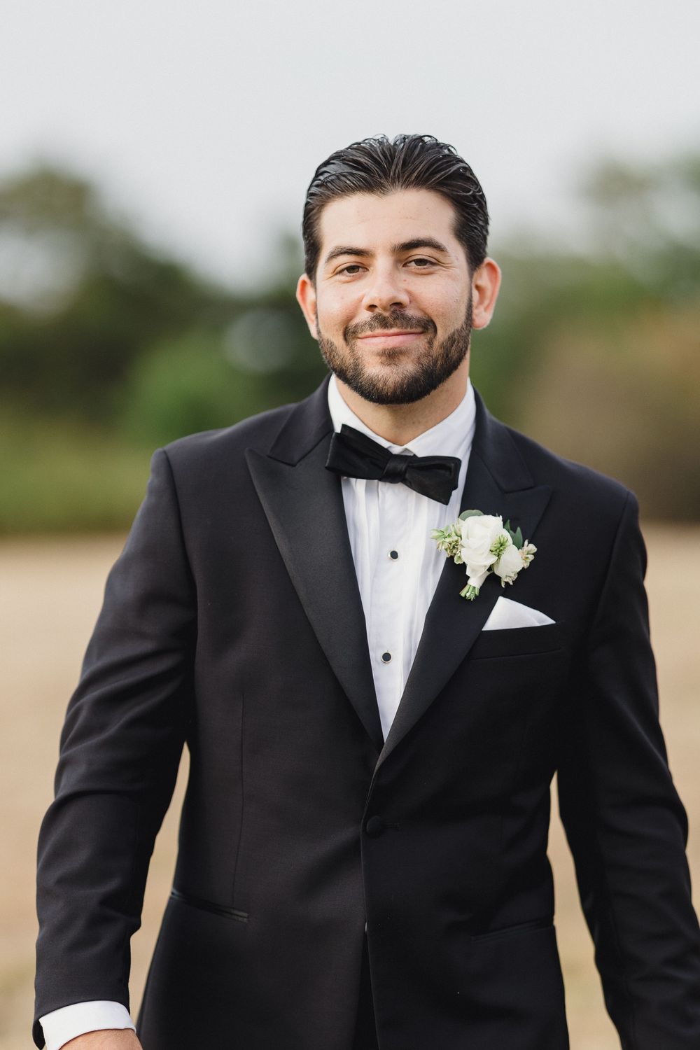 Dapper groom in black tuxedo with bow tie and white boutonniere smiling outdoors on wedding day. OceanCliff Newport RI Wedding