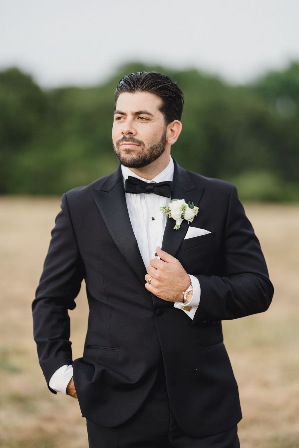 Groom in black tuxedo with bow tie and white boutonniere poses outdoors in a field on his wedding day. OceanCliff Newport RI Wedding
