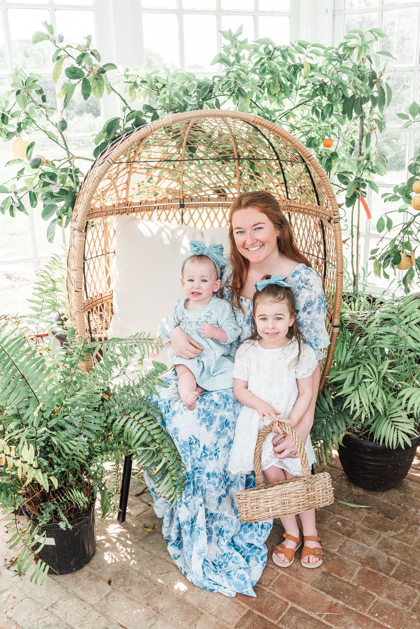 A sweet mother and two girls moment captured in a greenhouse setting with matching blue floral dresses and natural light streaming through.