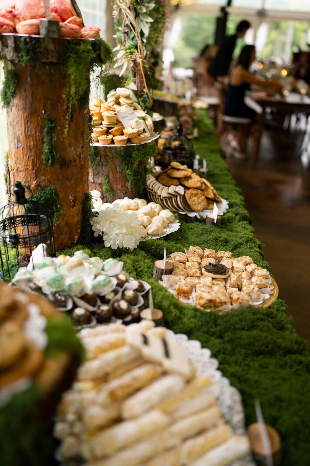 Rustic wedding dessert table with a moss runner, tree stump stands, pastries, cookies, and sweets displayed on platters.