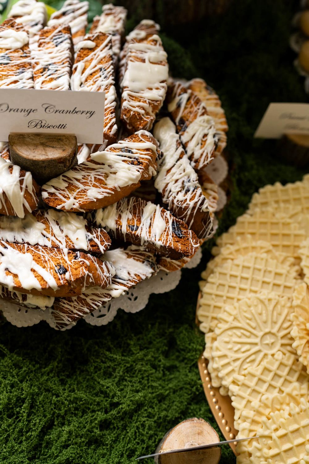 Orange cranberry biscotti drizzled with white icing arranged on a doily-lined tray beside pizzelle cookies on a moss-covered dessert table.