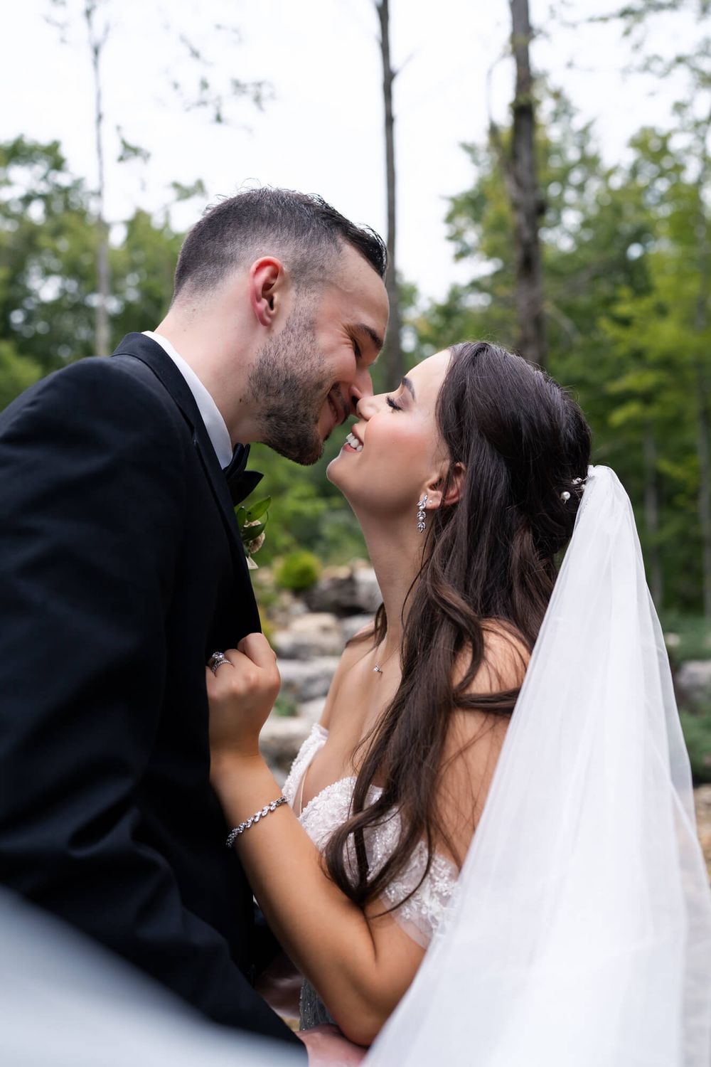 Smiling bride in a lace gown and long veil touches noses with her groom in a black suit during an outdoor woodland wedding portrait.