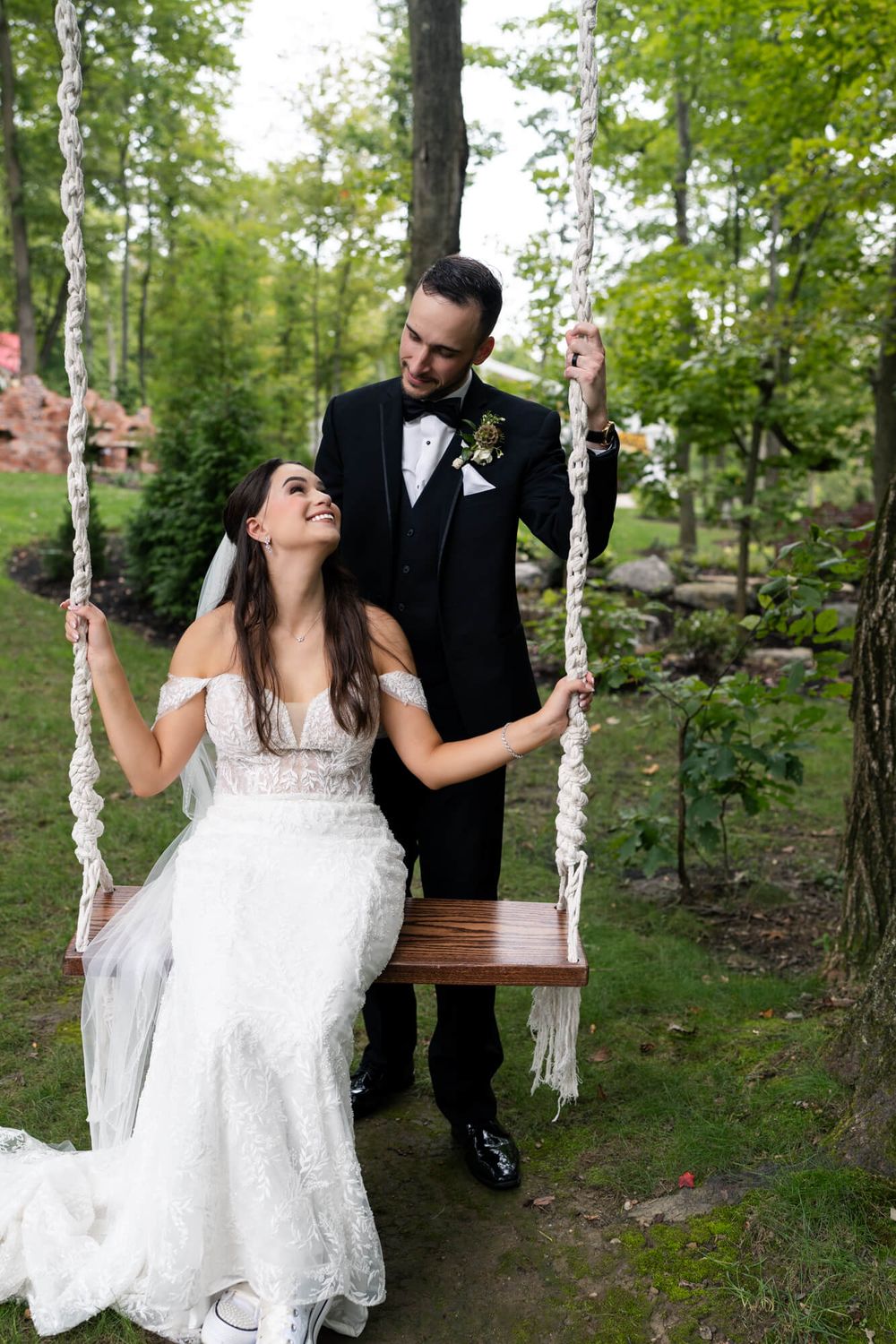 A bride in a lace wedding gown sits on a rope swing in a wooded garden as the groom in a black tuxedo smiles down at her.
