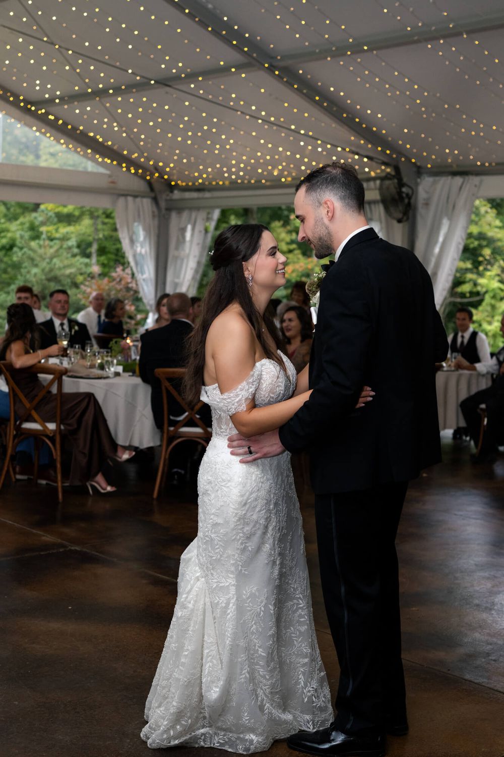 Bride in a lace off-shoulder gown and groom in a black suit share a first dance under a tented reception with string lights.