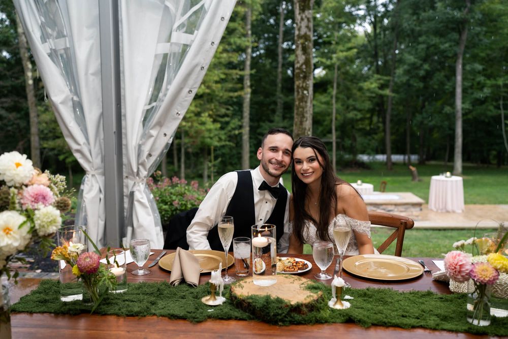 Newlywed couple smiles at a rustic reception table with floral centerpieces, wine glasses, and a moss runner in a sunlit garden.
