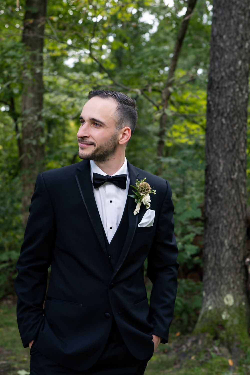 Groom in a black tuxedo with bow tie and floral boutonniere stands in a green forest, smiling and glancing to the side.