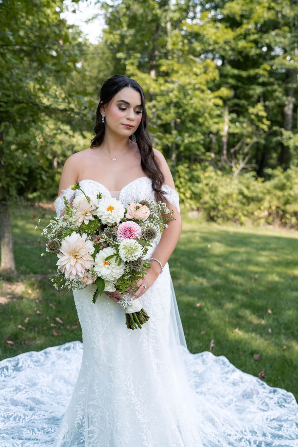 Bride in an off-shoulder lace wedding gown holds a pastel dahlia bouquet while standing on a sunlit lawn framed by green trees.