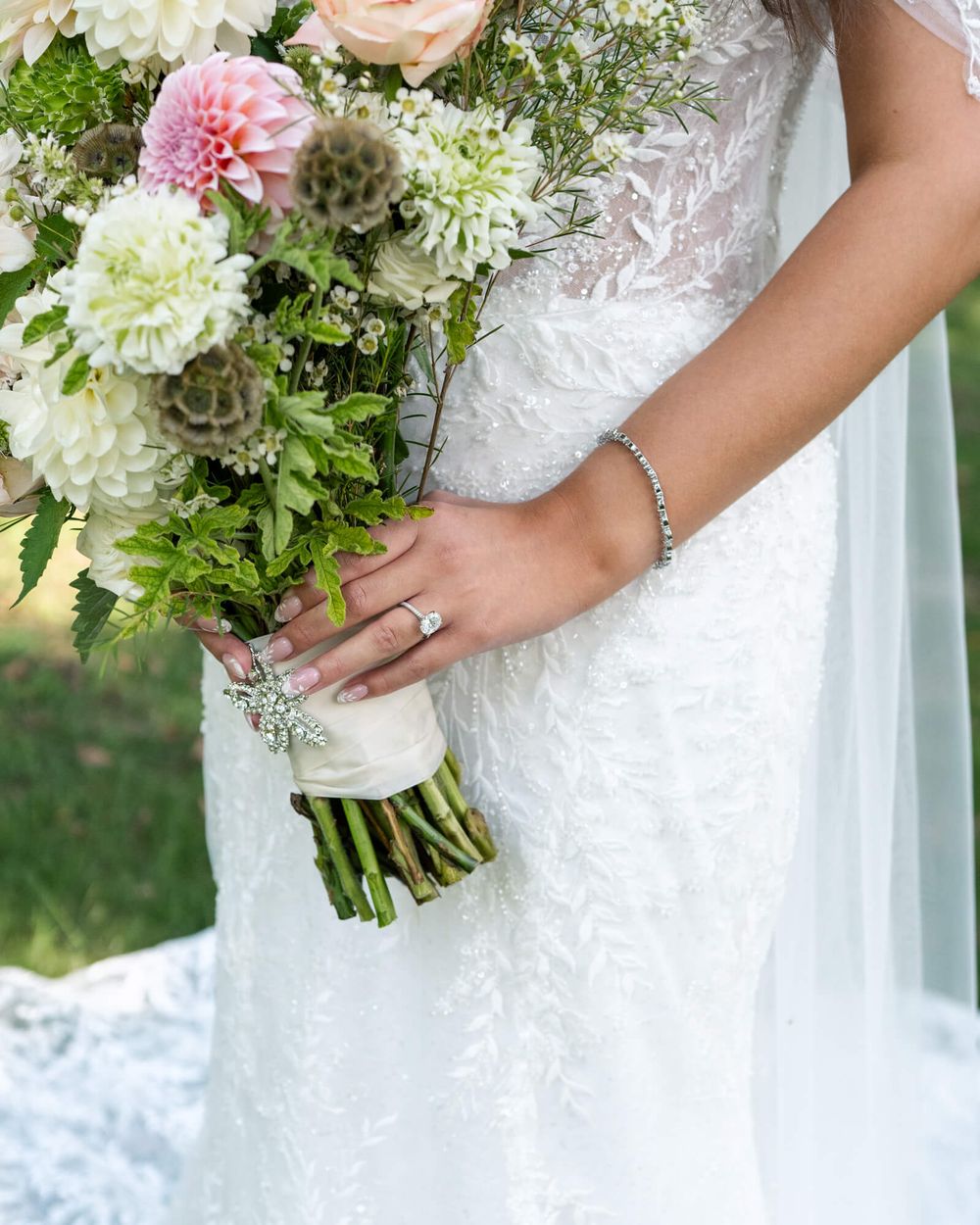 Bride in a lace wedding gown holds a bouquet of pink dahlias, white blooms and greenery wrapped in ivory ribbon.