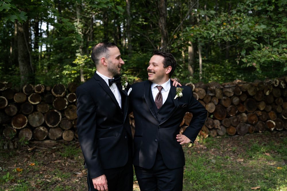 Two smiling grooms in dark suits and boutonnieres embrace in front of a stacked firewood wall in a sunny forest setting.