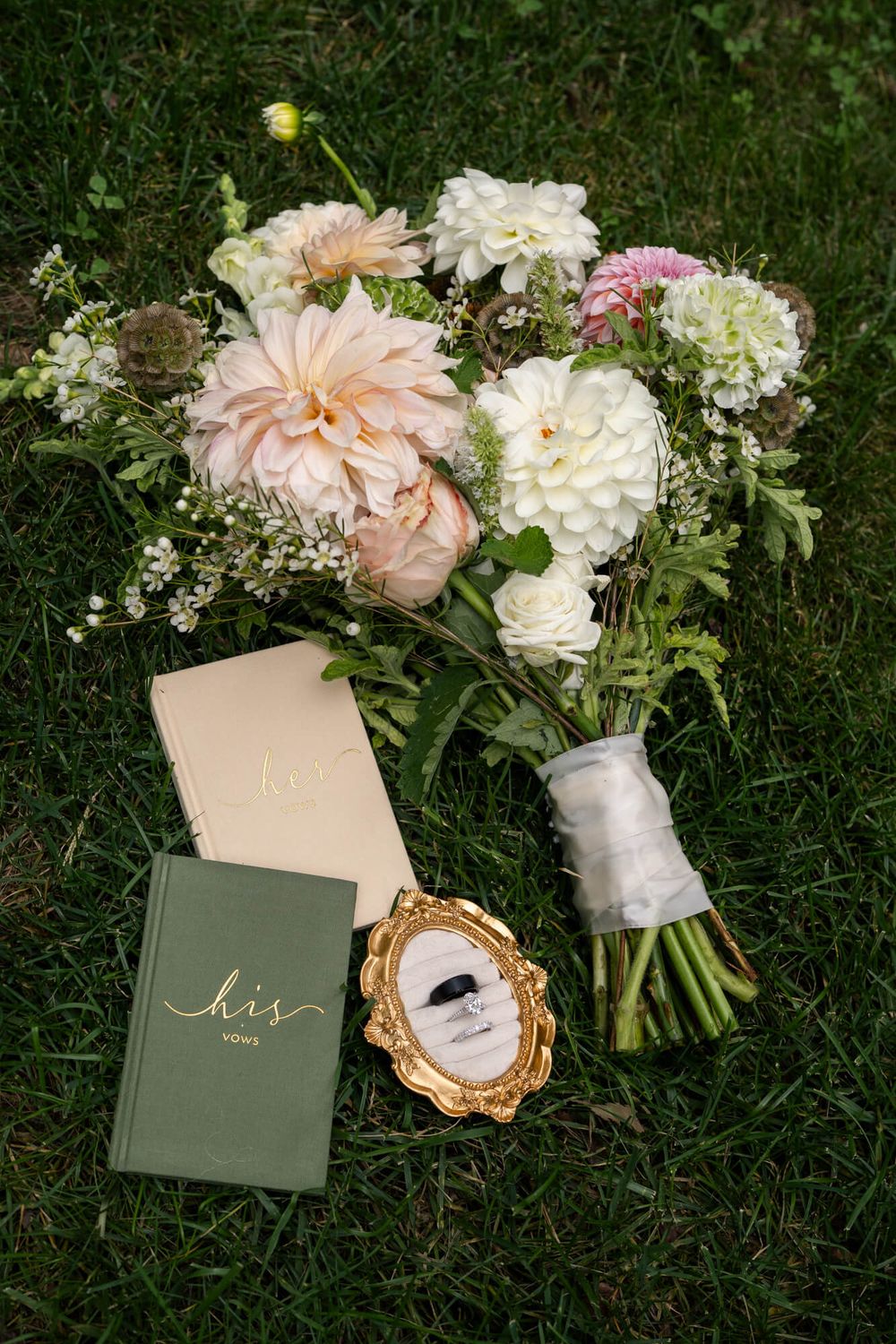 Wedding bouquet of blush dahlias and white blooms on grass beside his and hers vow books and a gold framed ring photo.