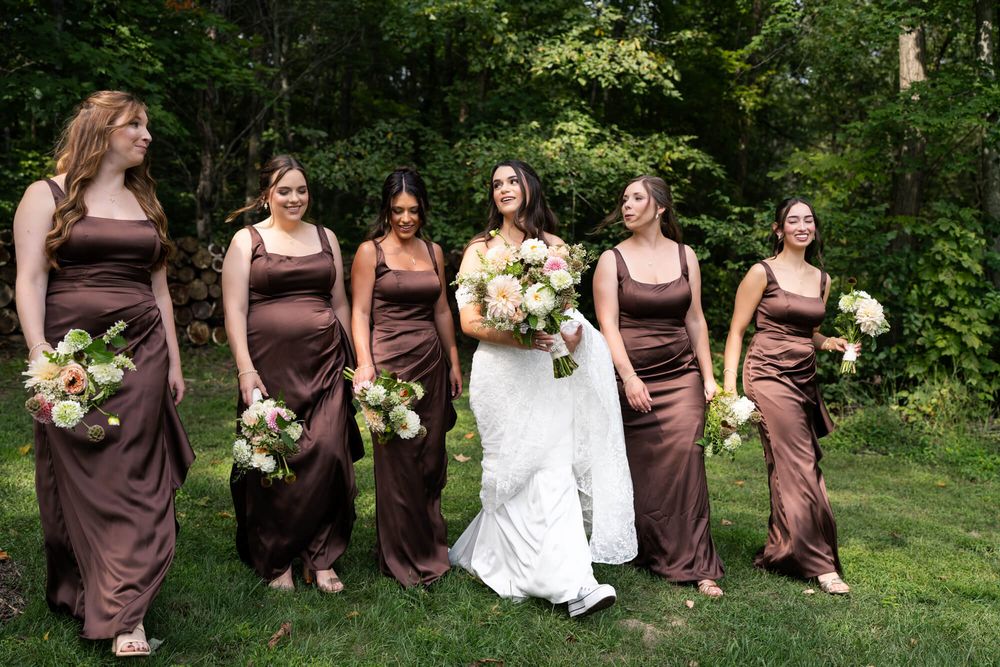 Bride in a white gown walks with five bridesmaids in brown dresses, all holding floral bouquets, across a green lawn with trees.