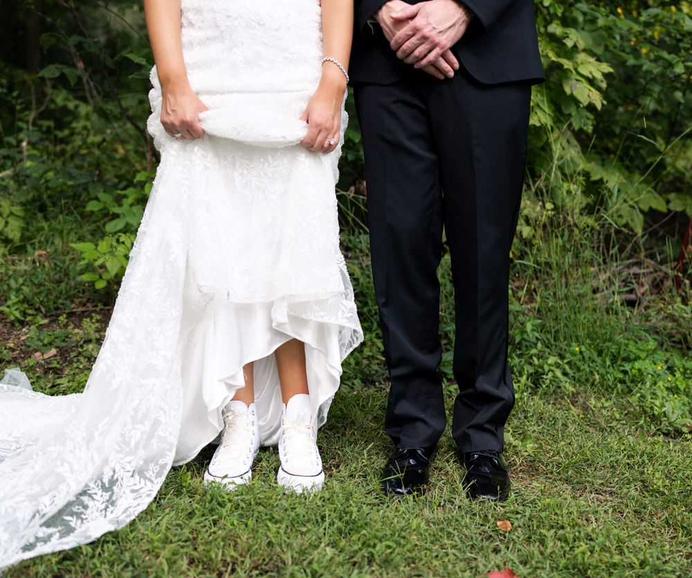 Bride in a lace wedding gown and white high-top sneakers stands on grass beside a groom in black pants and dress shoes.