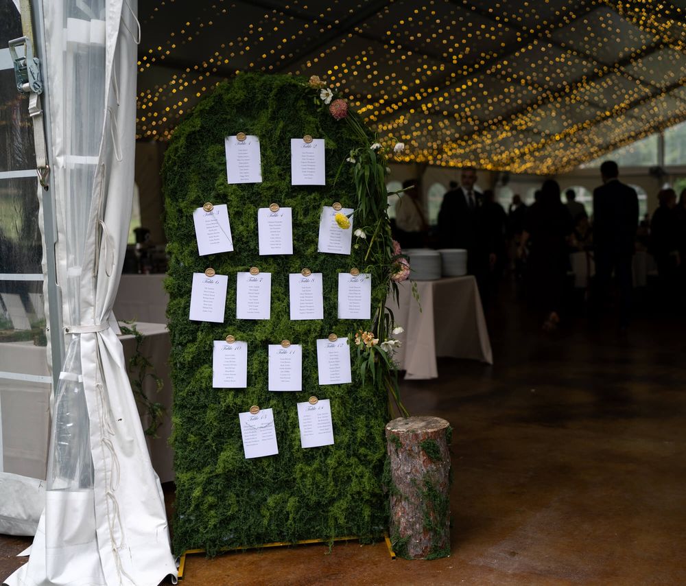 Wedding seating chart with white place cards on a green moss wall beside a tree stump under a tent with string lights.
