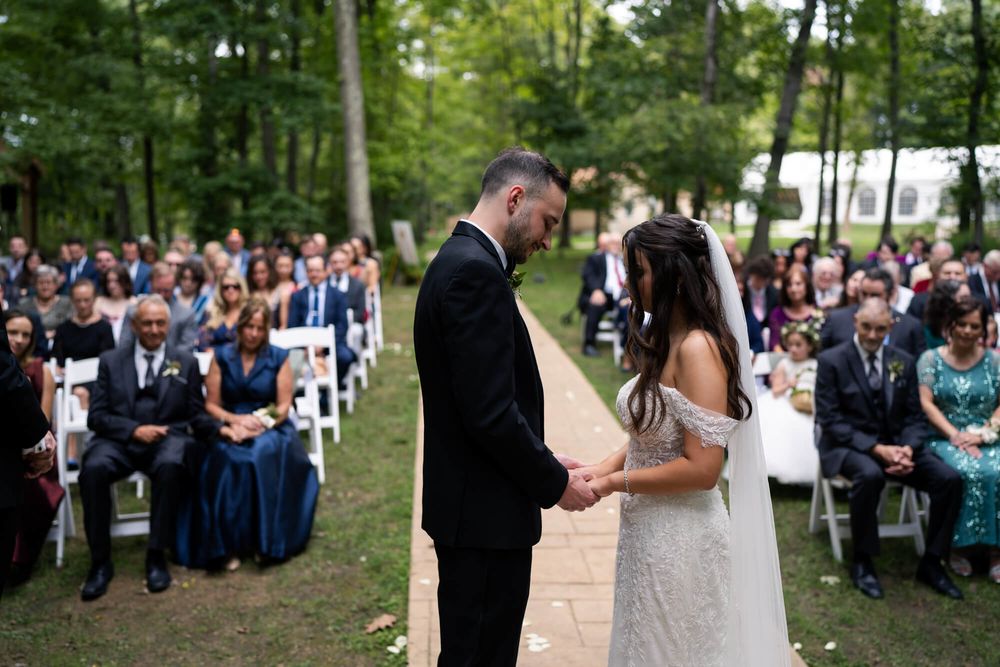 Bride and groom hold hands while exchanging vows during an outdoor wedding ceremony as seated guests watch among tall trees.