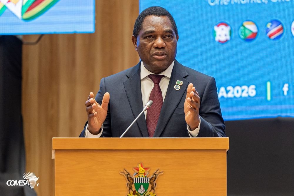 Speaker in a dark suit and maroon tie gestures while addressing a conference from a wooden podium with a coat of arms and COMESA logo.