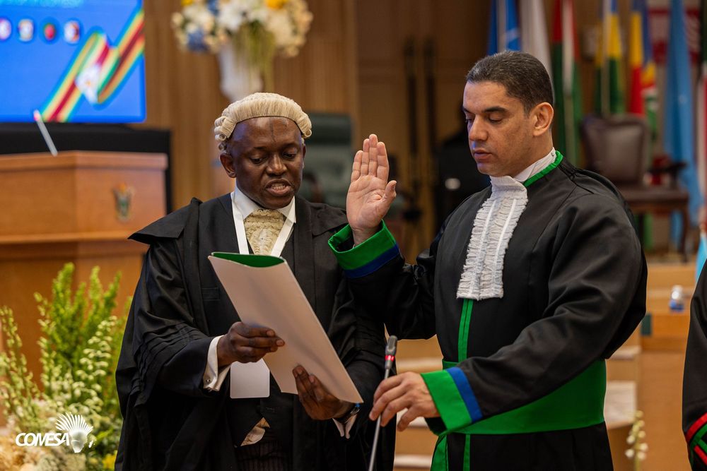 Judge in a white wig and black robe holds a document while a man in a green-trimmed robe raises his right hand during a swearing-in ceremony