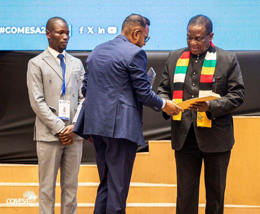 Three men on stage at a COMESA event, with one presenting a document to a man wearing a striped scarf in Zimbabwe flag colors.