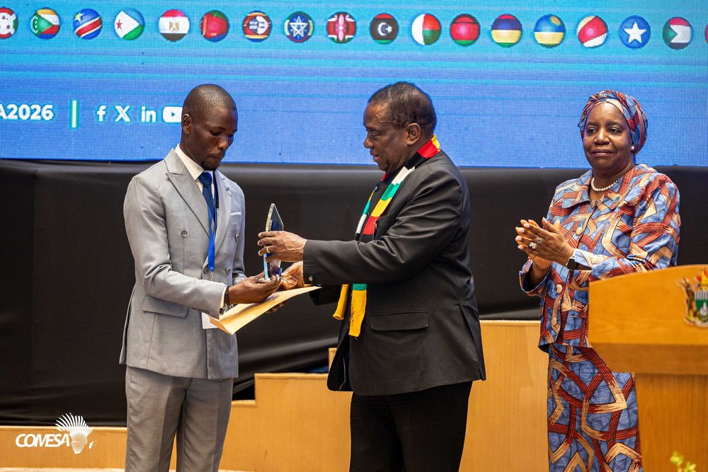 Man in a gray suit receives a glass award from a man in a dark jacket and striped scarf as a woman applauds at a COMESA event.