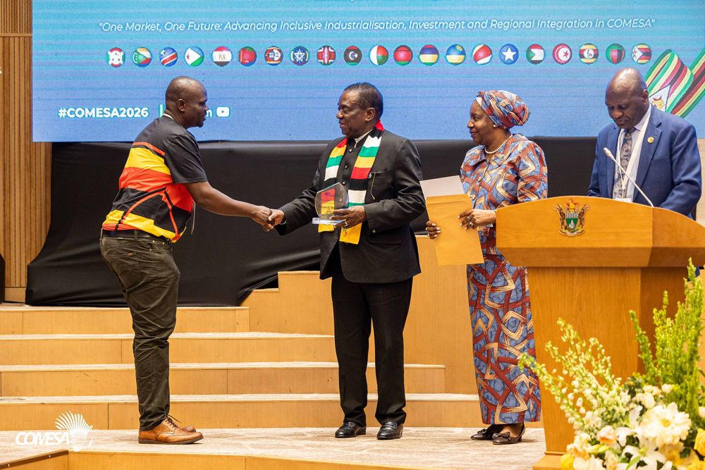 Two men in colorful striped scarves shake hands on stage at a COMESA 2026 event as a woman and an official at a podium look on.