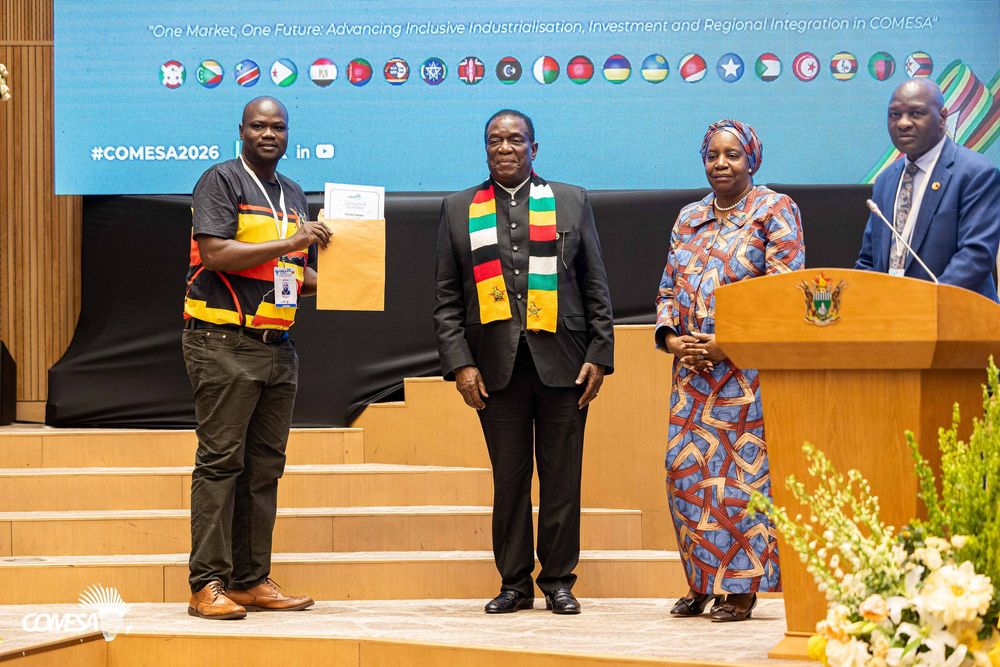 Three people pose on a stage beside a wooden podium with flowers at a COMESA 2026 event, one in a colorful striped scarf.