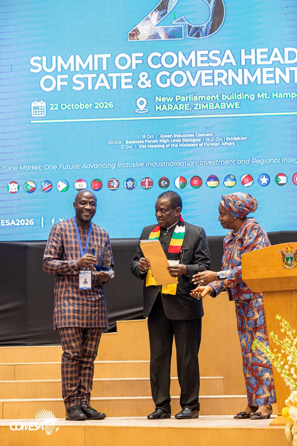 Three officials on stage at the COMESA Summit of Heads of State and Government in Harare, Zimbabwe, exchanging a document and award.