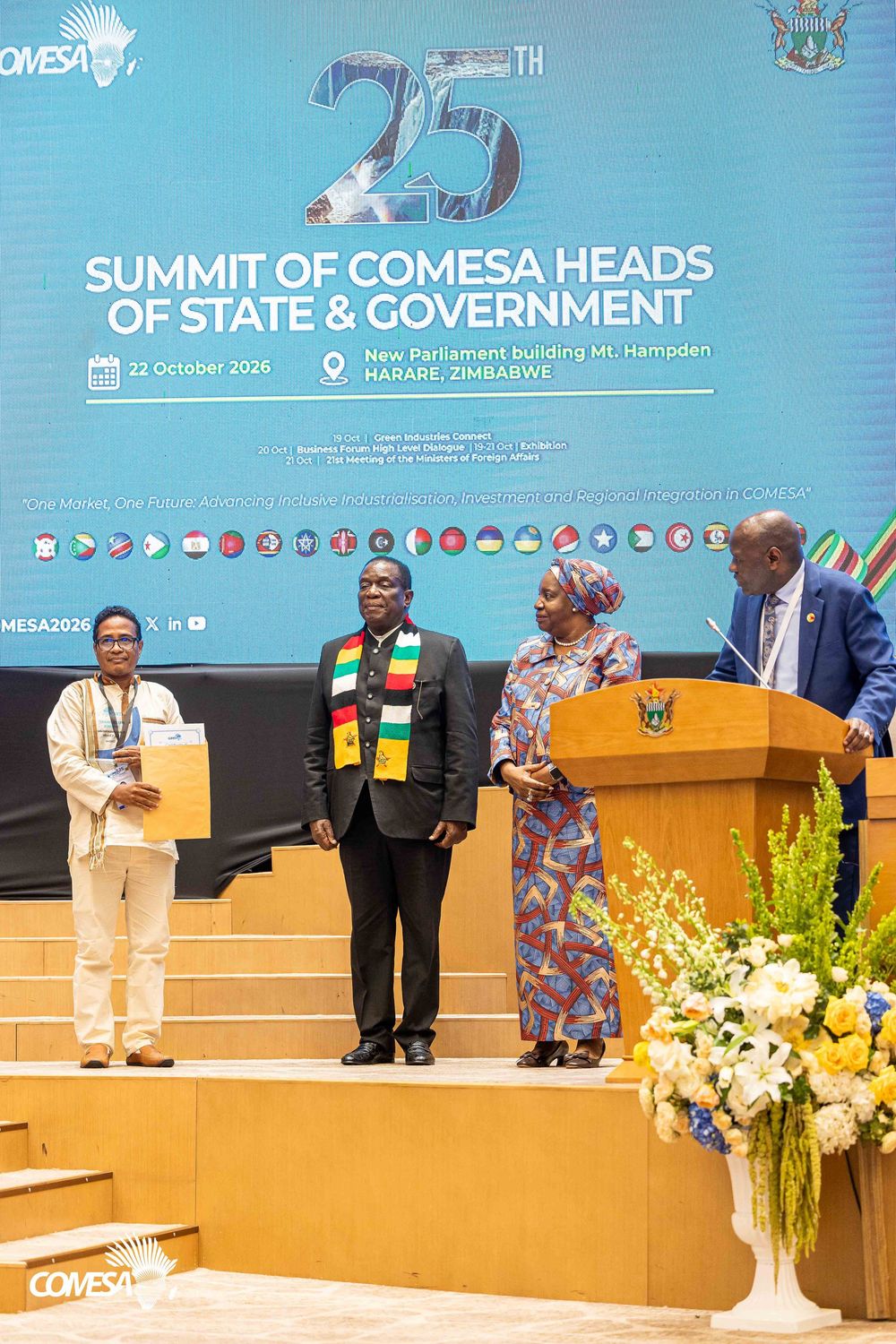 Officials on stage at the 25th COMESA Summit of Heads of State and Government in Harare, Zimbabwe, with a podium and flowers.
