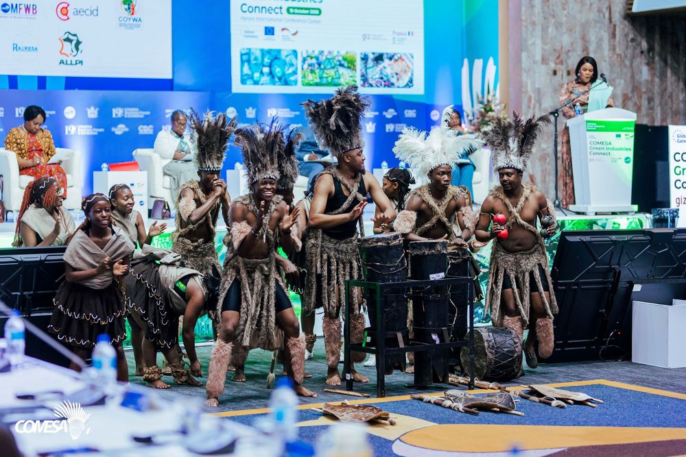 Performers in traditional feathered headdresses and fiber skirts dance and drum on stage at a conference while attendees watch from nearby.