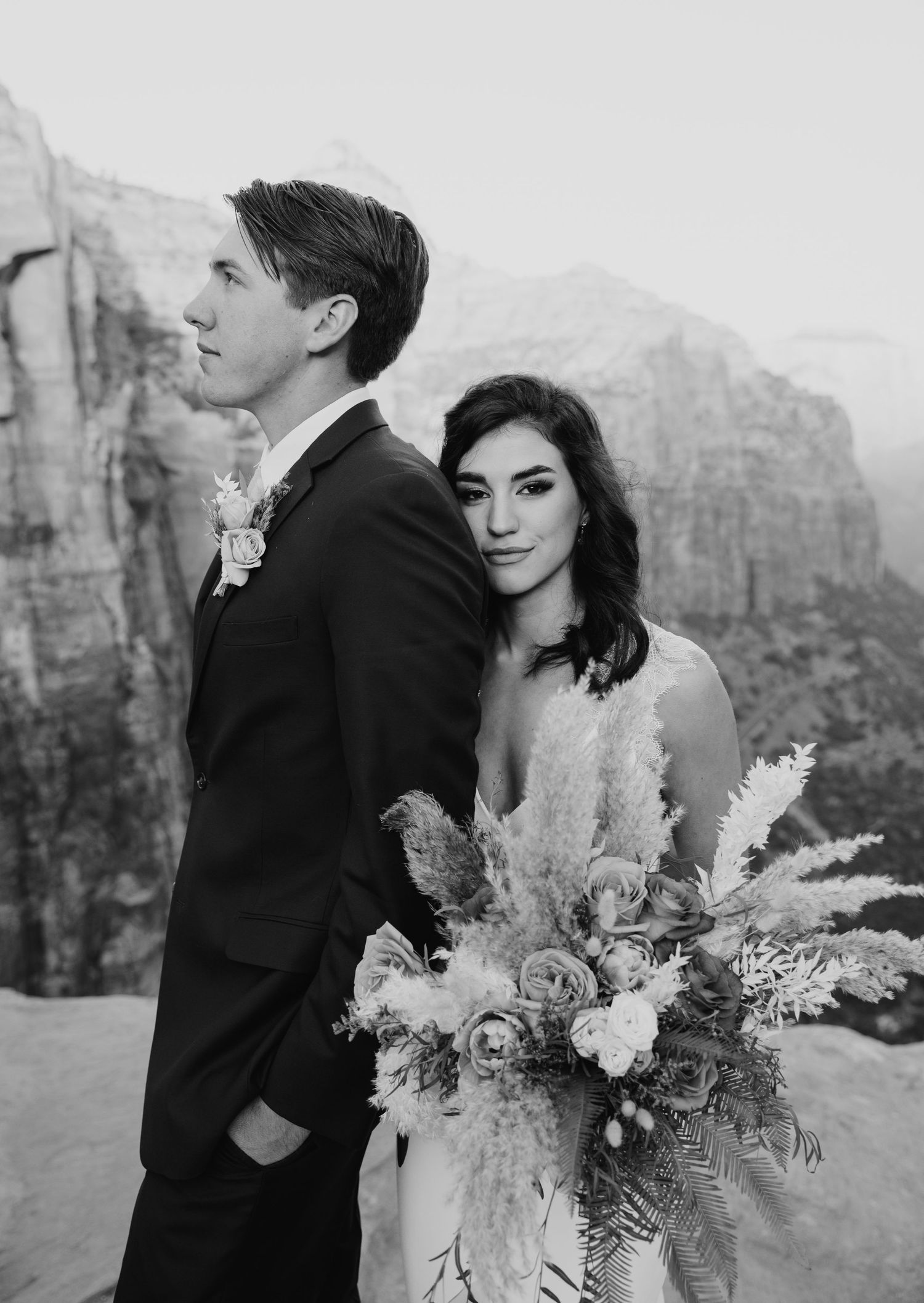 Black and white photo of a couple in wedding attire posing with a floral bouquet against a dramatic canyon backdrop.