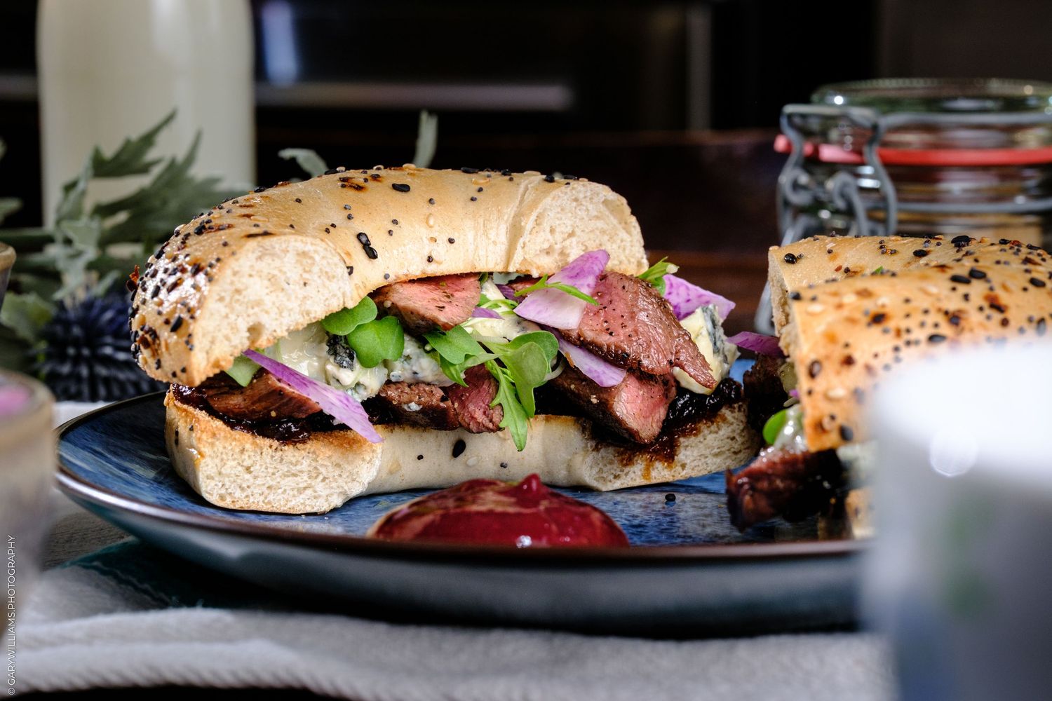 Gourmet steak sandwich on a seeded roll with arugula, red onion, and sauce served on a dark plate.