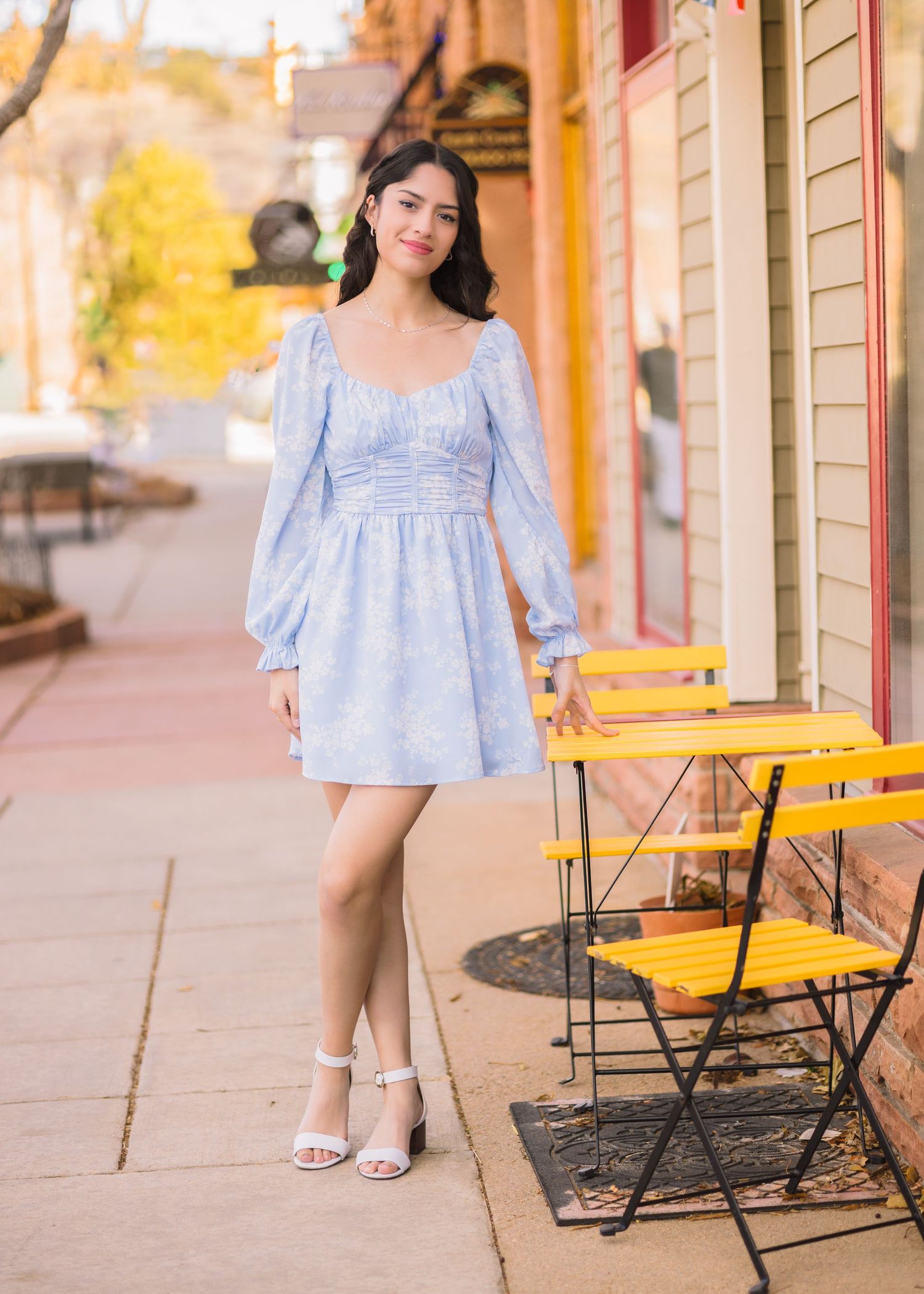 Young woman in a light blue floral mini dress with long sleeves poses on a sidewalk near a yellow cafe table.