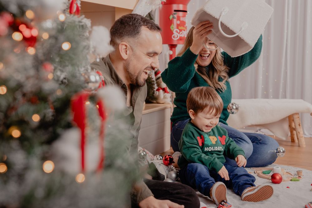 Family celebrating Christmas together, with parents and toddler opening gifts near a decorated tree in a cozy living room.