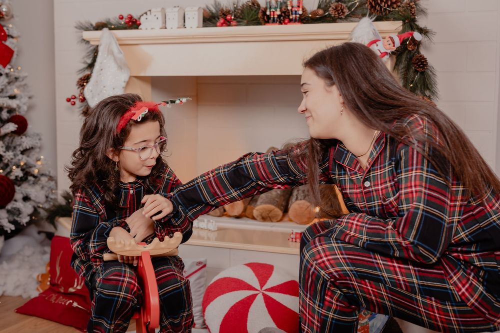 Mother and daughter in matching plaid Christmas pajamas sharing a festive moment by a decorated fireplace.
